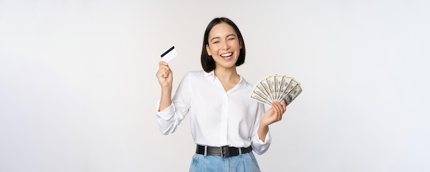 Happy korean woman holding credit card and money dollars smiling and laughing posing against white studio background