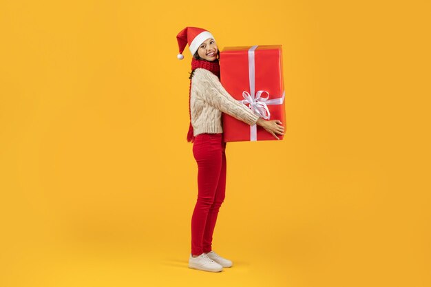 Happy lady in christmas attire holds big holiday present studio