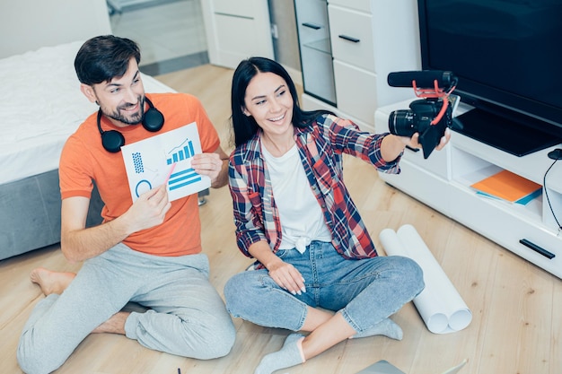 Happy lady holding a camera with a microphone and recording a smiling man with a graphic chart on a paper