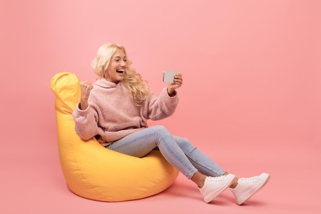 Happy lady holding smartphone and gesturing yes in joy reading good news sitting in beanbag chair