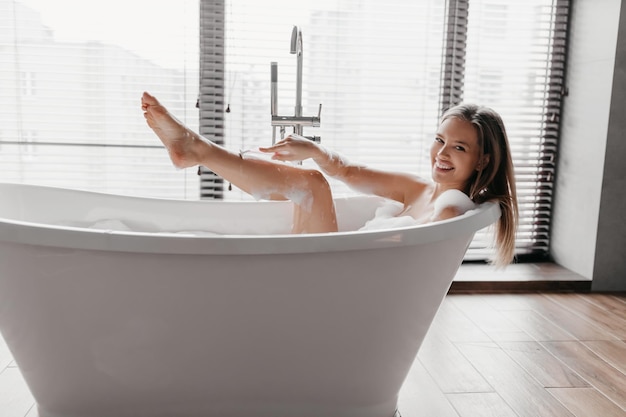 제모를 위해 안전 면도기를 사용하여 욕조에서 휴식을 취하고 다리를 면도하는 행복한 여성