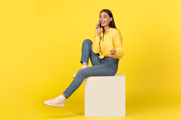 Happy lady talking on cellphone sitting on cube yellow background