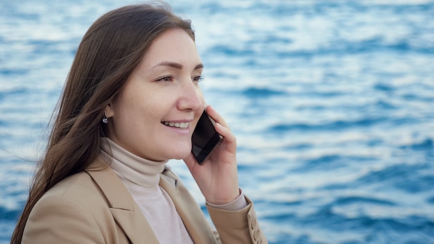 幸せな女性は、ぼやけた深い青色の波打つ海の極端なクローズビューに対して海岸に立っているスマートフォンで話します
