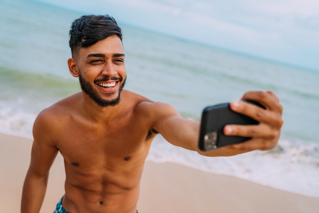 Felice uomo latino-americano che fa selfie sfondo spiaggia, soleggiati colori estivi