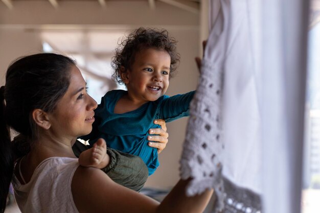 Happy Latin American mom 30 and her son 2 is lifted in arms and look out the window while playing at home Mother39s Day concept single mother