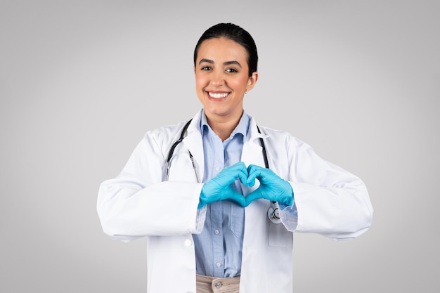 Happy latin lady doctor showing heart gesture with hands and smiling posing on gray studio