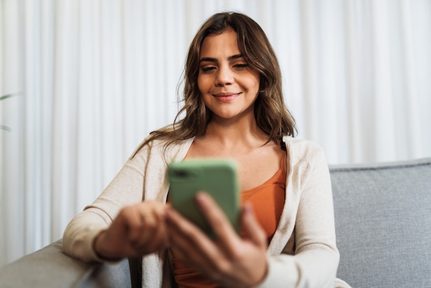 행복한 라틴 여성은 휴대전화를 통해 소셜 미디어에서 좋은 소식을 공유하는 방 포즈로 집에서 혼자 휴식을 취합니다. 웃고 있는 여성은 전화 앱에서 온라인으로 주말 주문 상품 음식을 즐깁니다.