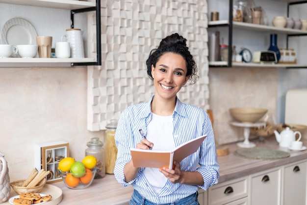 Happy latin woman holding cooking book writing recipe for delicious dinner standing in kitchen and