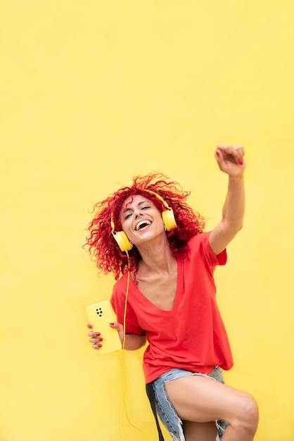 赤いシャツを着た黄色の背景の上にスマートフォンを持ち、黄色のヘッドフォンで音楽を聴き、踊るアフロの赤い髪を持つ幸せなラテン女性