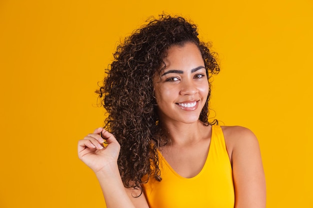 Happy laughing African American woman with her curly hair on yellow background. Laughing curly woman in yellow outfit touching her hair and looking at camera.