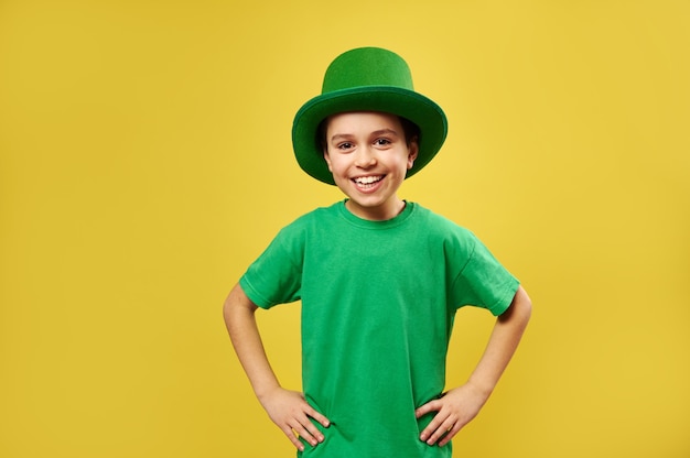 Happy laughing boy wearing leprechaun green hat poses to the camera on a yellow surface