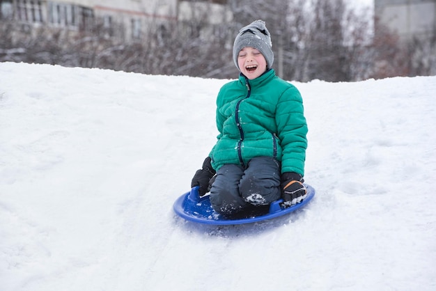 Happy laughing boy with closed eyes slides down the hill on snow saucer Seasonal concept Winter day