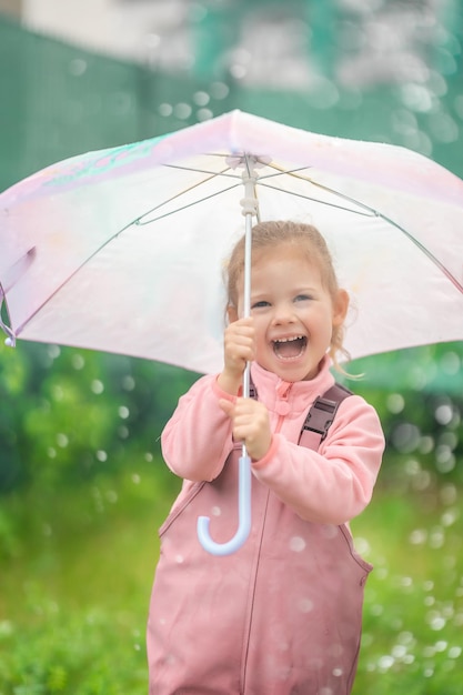 Happy laughing child girl 23 year old wearing waterproof clothes and holding pink umbrella have a fun on home backyard in rainy day