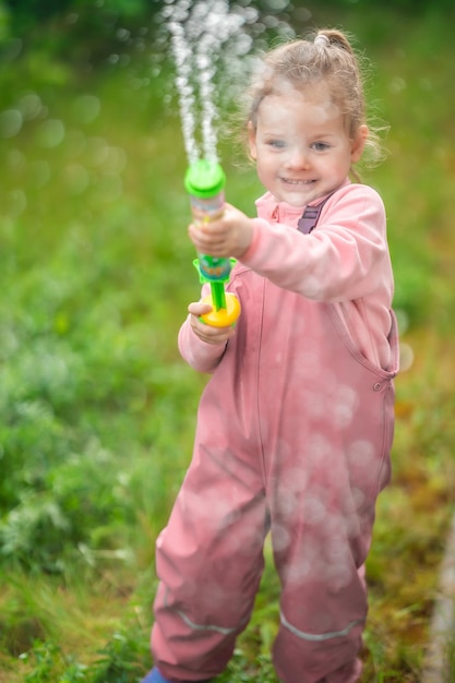 Счастливая смеющаяся годовалая девочка в водонепроницаемой одежде развлекается с водяным пистолетом на спине дома