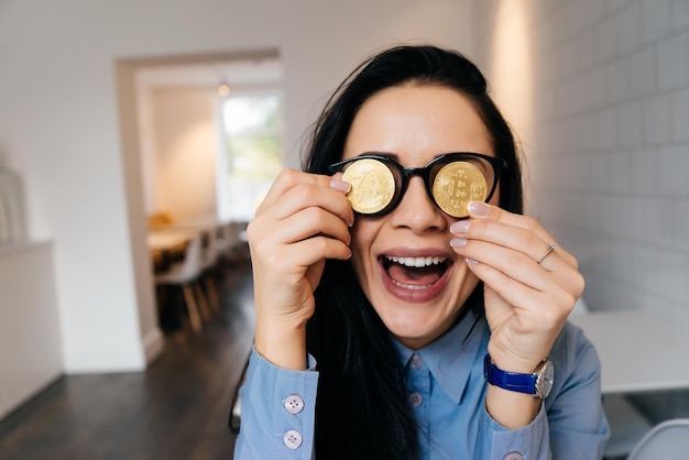 Happy laughing girl holding golden bitcoins by the eyes