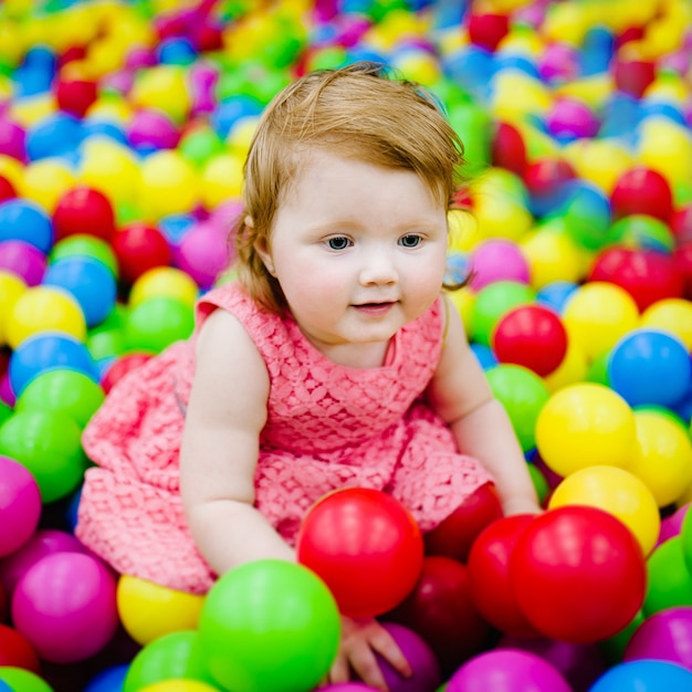 Happy laughing girl playing with toys, colorful balls in playground, ball pit, dry pool. Little cute child having fun in ball pit on birthday party in kids amusement park and play center indoor.