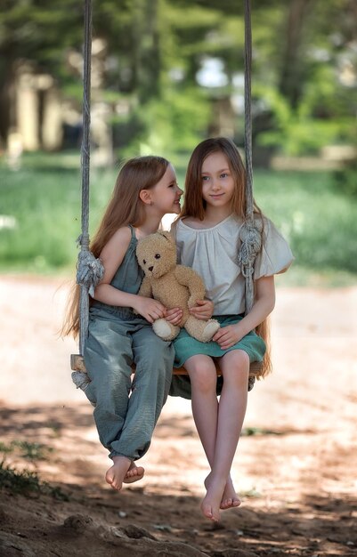 Happy laughing kids girls sisters with long hair enjoying a swing ride with a teddy bear toy on a sunny summer day