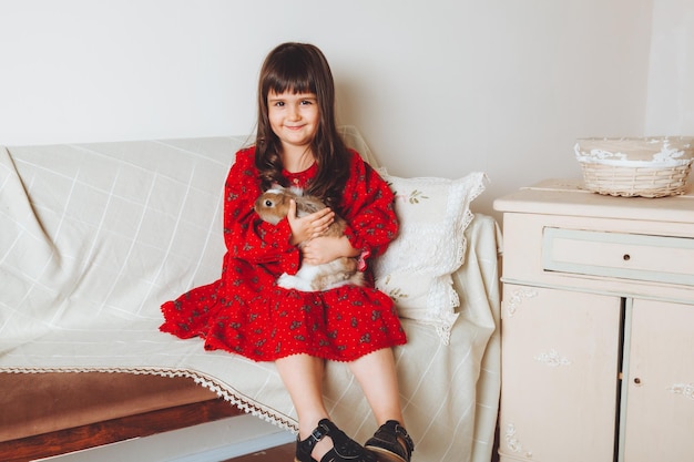 A happy laughing little girl in a red dress plays with a baby\
rabbit hugs a pet rabbit and learns to take care of the animal baby\
sofa at home