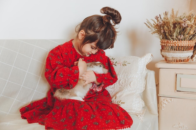 A happy laughing little girl in a red dress plays with a baby rabbit hugs a pet rabbit and learns to take care of the animal Baby sofa at home