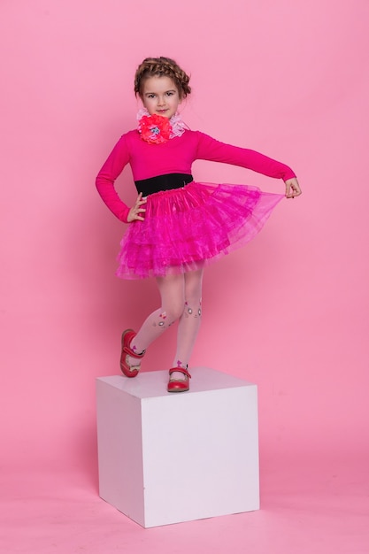ピンクの背景で踊る幸せな笑いの小さな長い髪の少女。ピンクの背景に白い立方体の少女