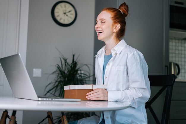 Happy laughing redhead young woman opening gift box with\
present during video call on laptop sitting at desk. concept of\
leisure activity red-haired female at home during\
self-isolation.