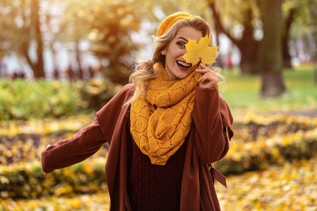 幸せな笑いの女性は、秋の葉を手に黄色いニットベレー帽の黄色い葉で目を隠し、黄色い庭や公園に落ちる