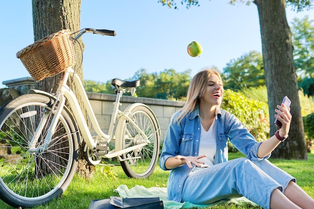 Happy laughing young woman sitting on the grass tossing juggling apple making video call on smartphone Active healthy beautiful woman with bike on park background