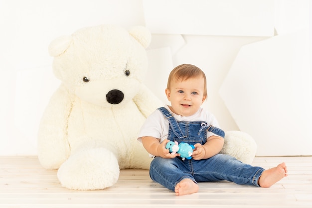 A happy lbaby boy six months old in a knitted warm jacket and blue jeans crawls at home near a large Teddy bear