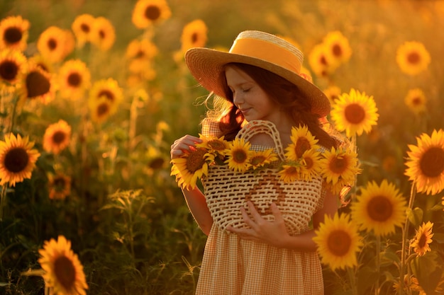 ヒマワリの畑に帽子をかぶった美しい赤い髪の少女の夏のポートレートでの幸せな生活、夕日の光の中で彼女の手の中の花