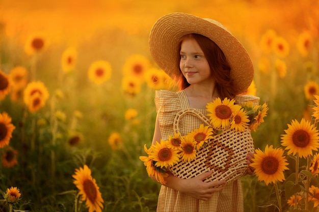 ヒマワリの畑に帽子をかぶった美しい赤い髪の少女の夏のポートレートでの幸せな生活、夕日の光の中で彼女の手の中の花