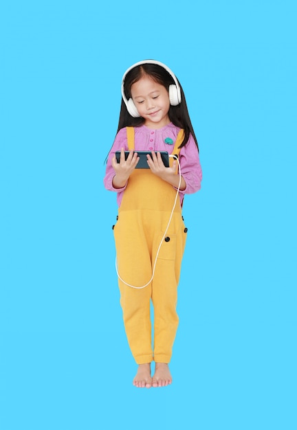 Happy little Asian child girl in pink-yellow dungarees with headphones to enjoys listening music 