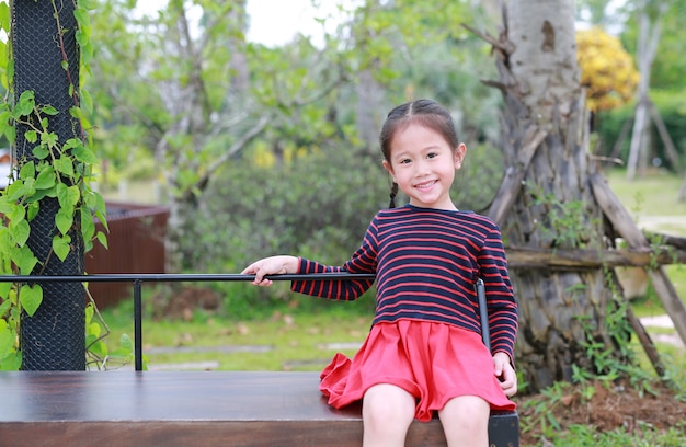 Happy little Asian child girl sitting on wood bench in the public garden.