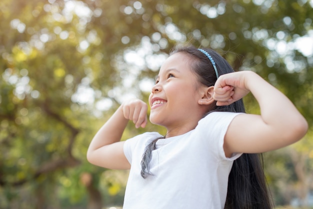 幸せな小さなアジアの女の子の子供が大きな笑顔で前歯を見せて立っています。腕の筋肉が誇らしげに笑っているのを見せています。