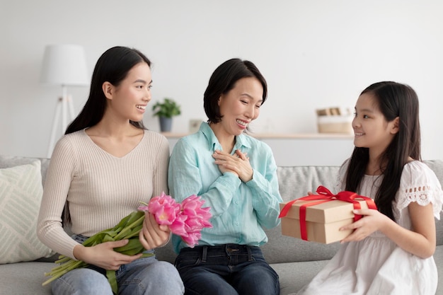 Happy little asian girl giving her mother and grandmother wrapped gift and flowers greeting them