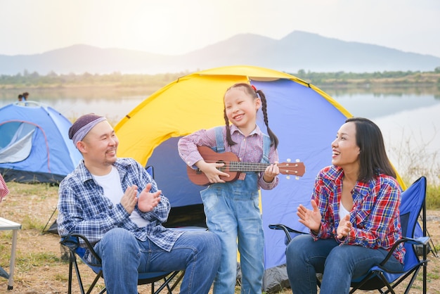 Felice piccola ragazza asiatica che suona l'ukulele e i suoi genitori che battono le mani al campeggio