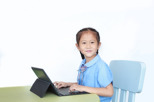 Счастливая маленькая азиатская девушка используя портативный компьютер для того чтобы сделать домашнюю работу на столе.