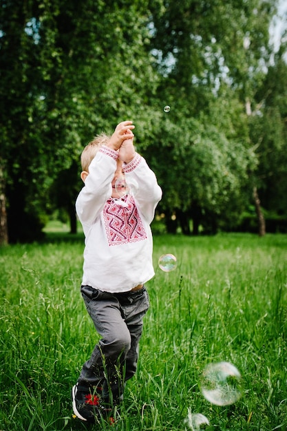 Happy little baby boy run in embroidered shirt on the background of nature upper half Close up Catch inflatable bubbles