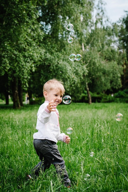 Happy little baby boy running in embroidered shirt on the background of nature full length looking sideways Close up Catch inflatable bubbles