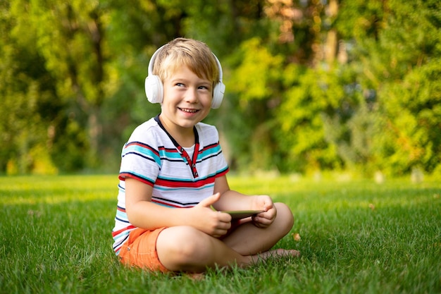 Счастливый маленький мальчик в беспроводных наушниках держит планшетный компьютер на улице в летнем парке, улыбаясь