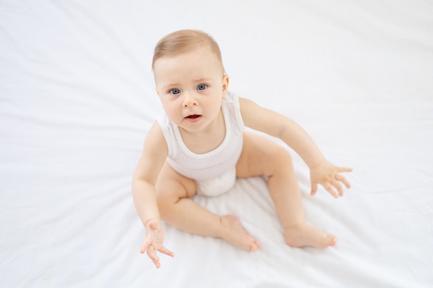 Happy little baby girl or boy in a white bodice on the bed in the bedroom looking up baby with blue eyes