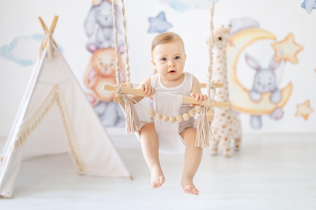 a happy little baby girl or boy in a white body swings on a swing in the nursery the kid has fun smiling or laughing