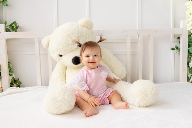 Happy little baby girl six months old sitting on a white bed in pink clothes, with a big Teddy bear and smiling