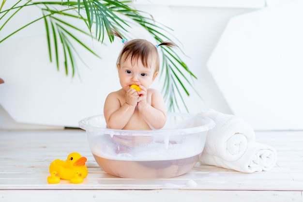 Happy little baby girl washes in a basin with foam and water in a bright room at home and plays with a yellow rubber duck