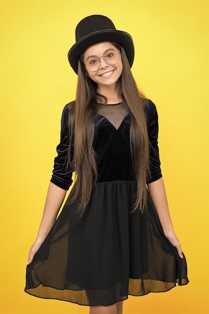 Happy little baby girl wear black party dress and hat on long brunette hair with beauty look yellow background salon