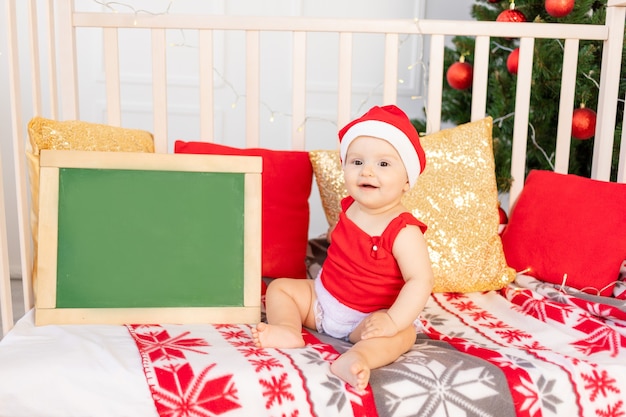 Happy little baby in a Santa hat sitting in a crib by the Christmas tree