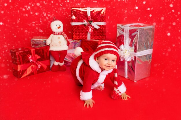 Happy little baby six months old in Santa costume sitting near gifts on red isolated photophone, space for text, new year and holiday concept