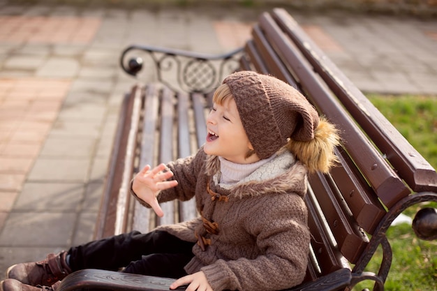 a happy little blond boy in knitted brown clothes is sitting on a park bench