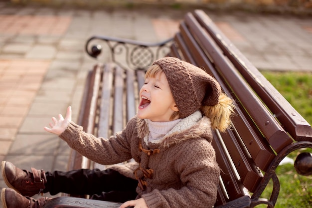 a happy little blond boy in knitted brown clothes is sitting on a park bench