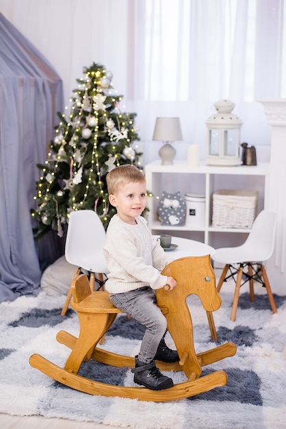 Happy Little blond boy rides a wooden rocking horse next to a Christmas tree with garlands