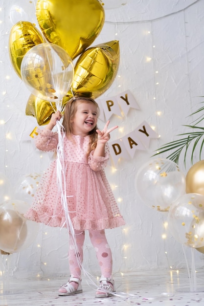 Happy little blond girl in pink dress holding balloons and celebrating her birthday